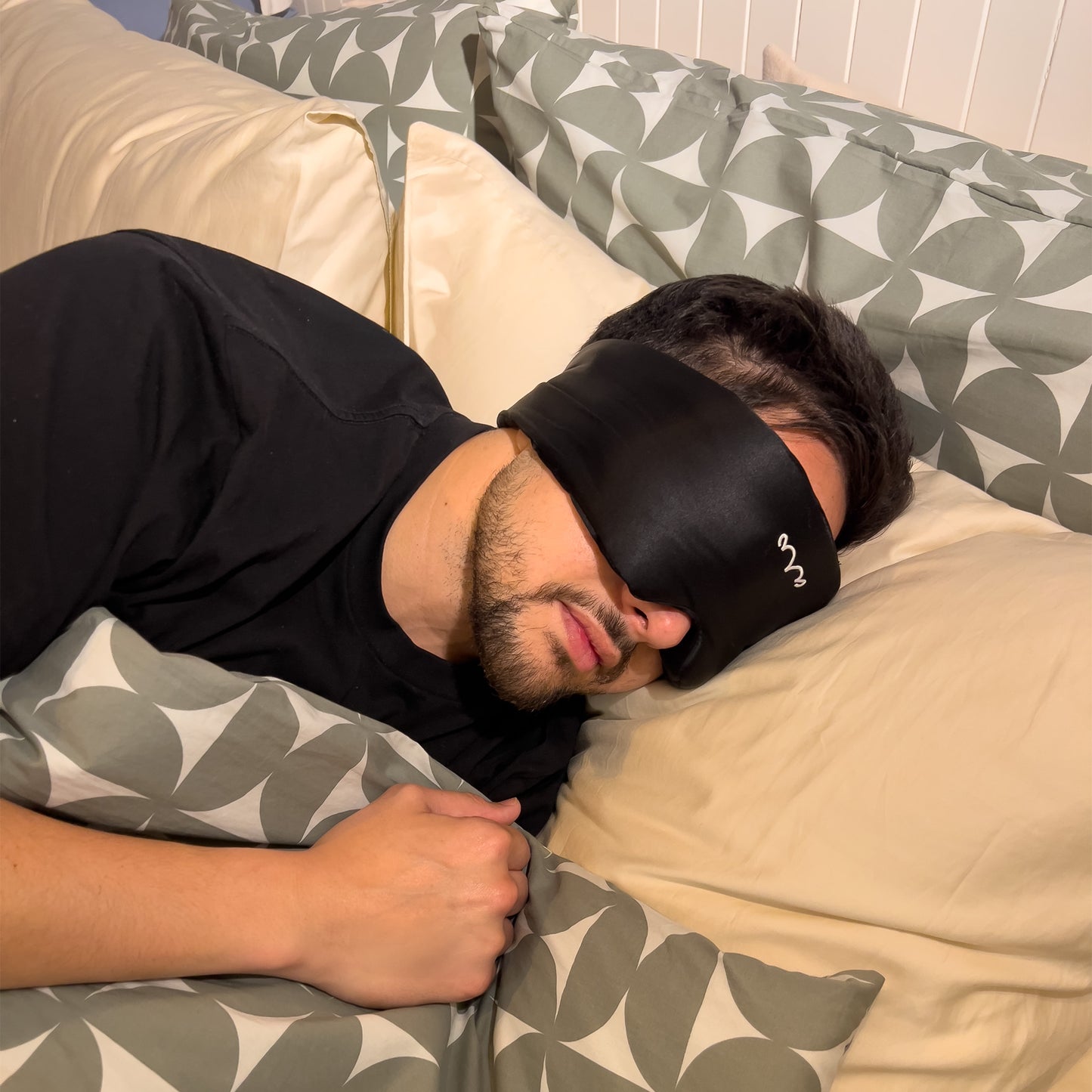 A photo of a young man lying in bed wearing the black MOE MANA 25 momme Mulberry Silk Sleep Eye Mask